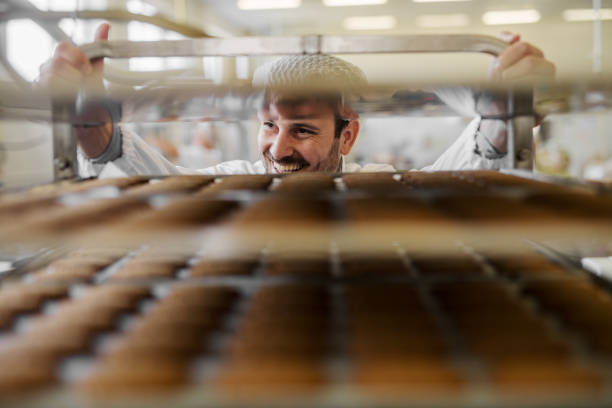 photo de l’homme de boulanger professionnel de mâles en uniforme de travail blanc. debout devant les étagères pleines avec biscuits frais. - baking food bread bakery photos et images de collection