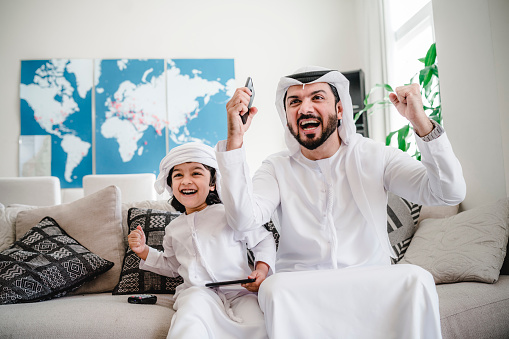 Arab dad and son playing with video games at home. Simple two colours world map with pins in the background.