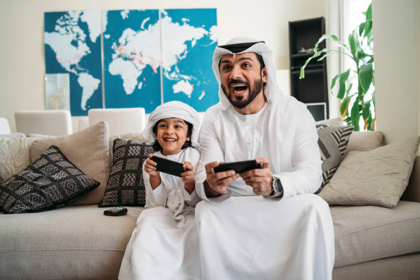Arab dad and son playing with video games at home Arab dad and son playing with video games at home. Simple two colours world map with pins in the background. arabian peninsula photos stock pictures, royalty-free photos & images