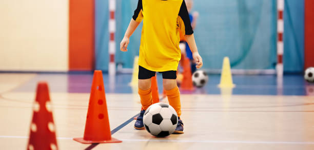 Football futsal training for children. Soccer training dribbling cone drill. Indoor soccer young player with a soccer ball in a sports hall. Player in orange uniform. Sport background Football futsal training for children. Soccer training dribbling cone drill. Indoor soccer young player with a soccer ball in a sports hall. Player in orange uniform. Sport background cone shape photos stock pictures, royalty-free photos & images