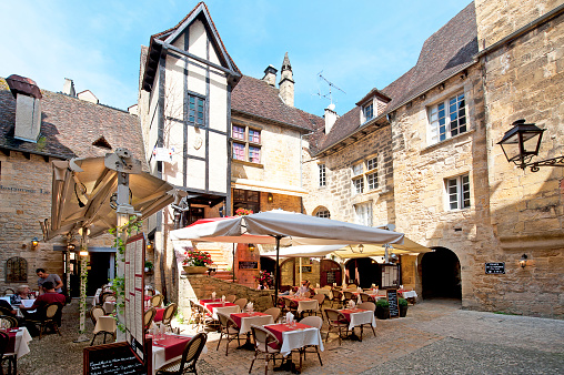 Sarlat-la-Canéda, Dordogne, Nouvelle-Aquitaine, France. Outdoor pavement café restaurant with parasol covered tables and chairs near the town square, typical traditional architecture style of the homes and houses found in the backstreet alleys between buildings of timber frame and stone construction in the ancient medieval old town of Sarlat, in the department of Dordogne, southwest France