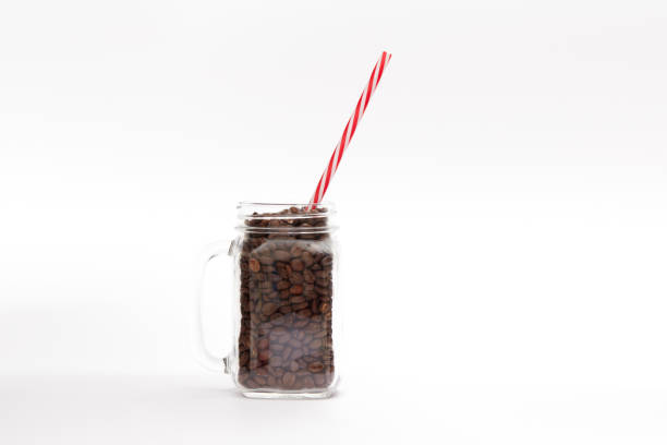 Coffee beans in a drinking glass or vintage jar with a straw on a white background with copy space