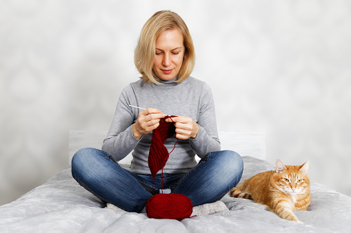 Woman sits on the bed and knitting. Nearby is a red cat. Grey blurred background.