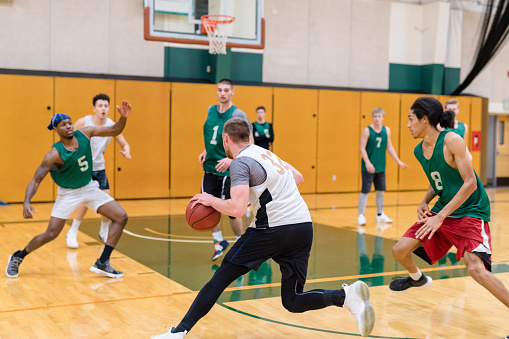 A men's college basketball team practices in the gym. They are scrimmaging and a player in the foreground is dribbling and getting ready to shoot.