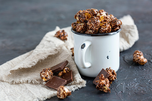 Chocolate caramel popcorn in enameled mug and pieces of milk chocolate on a textured dark background, selective focus.