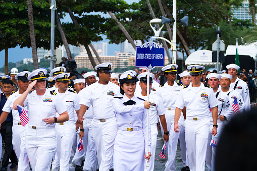 A group of US navy officers is walking in parade of asean fleet parade in Pattaya. Officers are greeting towards tribune. Along street people are standing.
