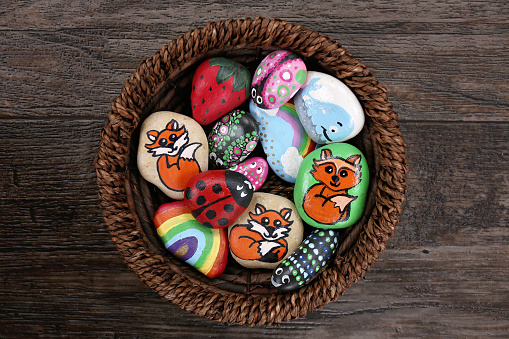 A collection of fun, handpainted, colorful cartoon rocks are together in a wicker basket, on a wooden plank background.
