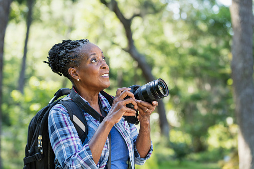 A senior African-American woman in her 70s enjoying the outdoors, hiking in a park, taking photographs. She is looking upward.