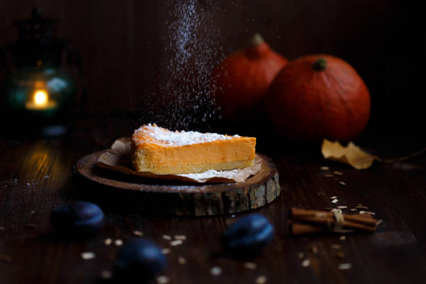 Female hand sprinkles powdered sugar on pumpkin cheesecake. Pumpkins, table lamp, foliage, vanilla on a wooden dark background. Autumn and winter cozy concept stock photo