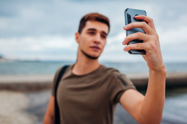 istock Young man taking selfie using phone on cloudy beach. Handsome sportive guy walking by pier 1049530768