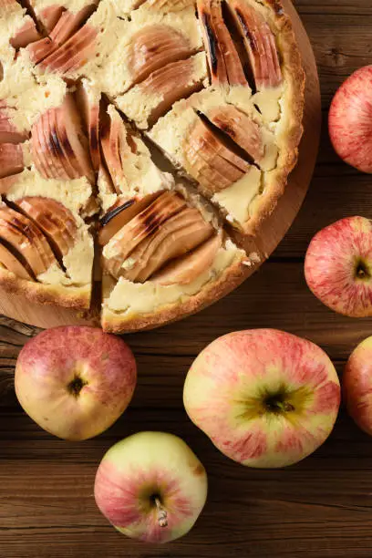 Piece of apple pie with cream filling served with raw organic apples on old wooden background closeup