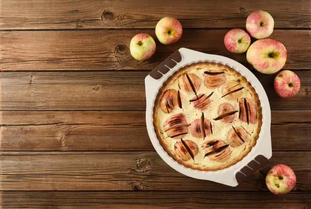 Autumn dessert. Homemade apple pie with cream filling in baking dish served with raw organic apples on old wooden background copyspace