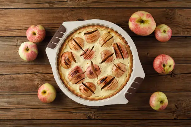 Yummy apple pie with cream filling in baking dish served with raw organic apples on old wooden background above view