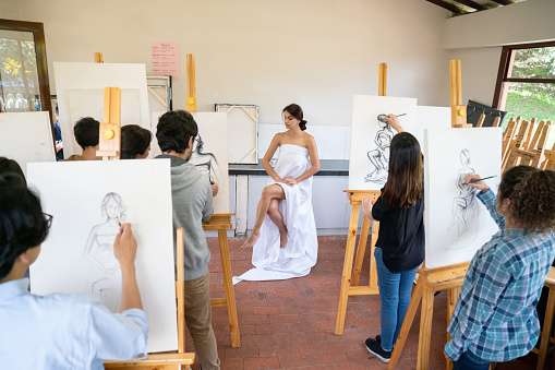 Group of people painting a live model in an art class using a canvas and an easel - education concepts