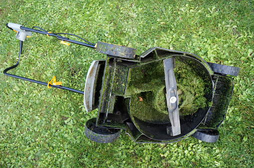 The electric lawn mower lies on the grass upside down with wheels. Inside, the entire mechanism is clogged with wet cut grass