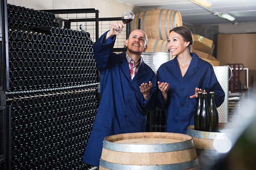 Two concentrated winery workers holding glass of wine in aging section of factory