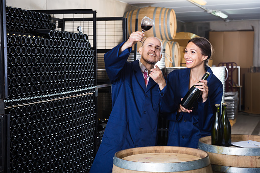Two friendly winery workers holding glass of wine in aging section of factory