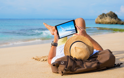 Unrecognizable male buying airline tickets online on tablet while relaxing on beach.