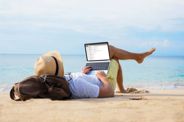 hombre leyendo correo electrónico en la computadora portátil mientras se relaja en la playa - trabajo freelance fotografías e imágenes de stock