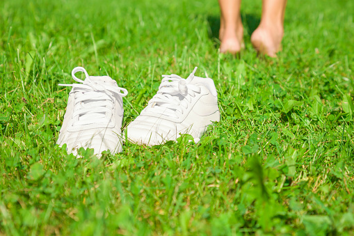 Young woman taked off white shoes after sport training. Walking on the fresh, green grass in sunny summer in the morning. Restful moment. Healthy lifestyle.