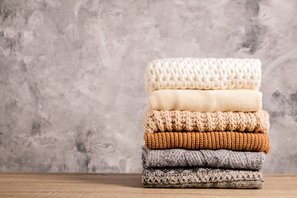 Minimalistic rustic composition with stacked vintage knitted easy chic oversized style sweaters, knitwear outfit. Bunch of knitted warm pastel color sweaters with different knitting patterns folded in stack on brown wooden table, grunged concrete wall background. Fall winter season knitwear. Close up, copy space sweater stock pictures, royalty-free photos & images