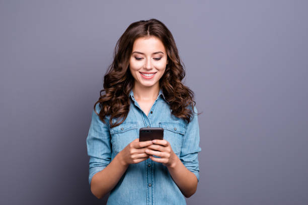 conteúdo na moda alegre bom bonita adorável adorável atraente menina morena com cabelos ondulados em camisa jeans casual, digitar no telefone, isolado sobre fundo cinza - girl - fotografias e filmes do acervo