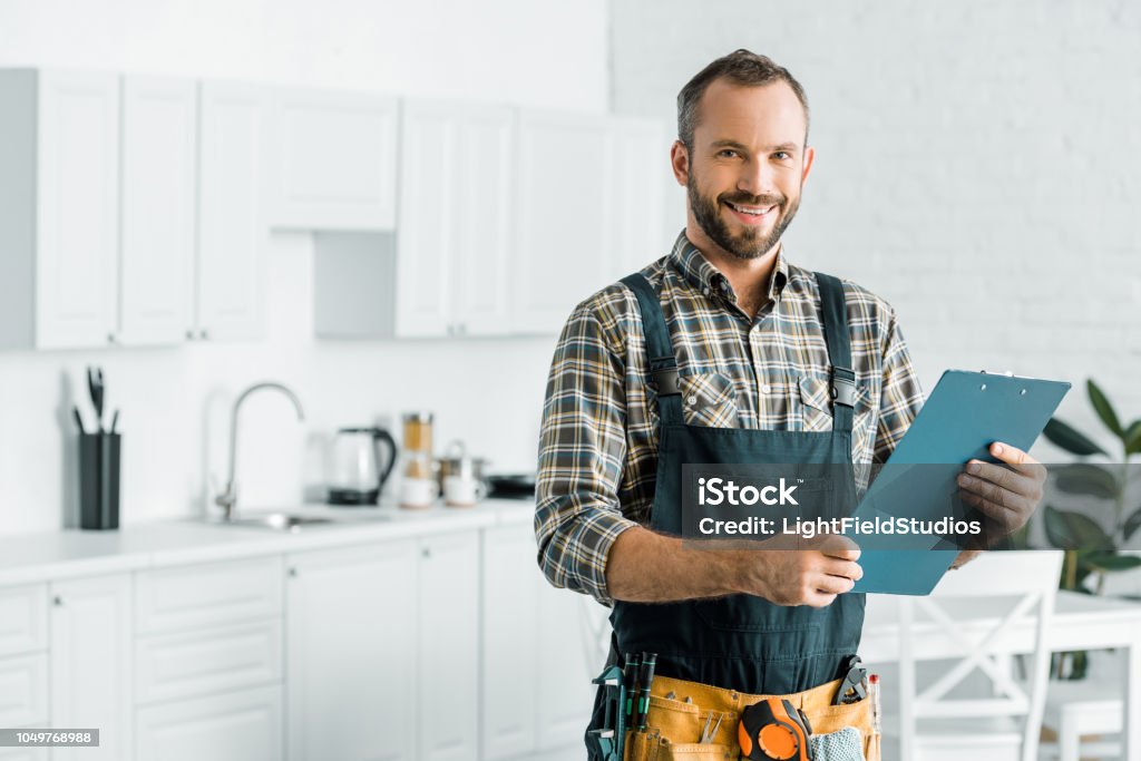 smiling handsome plumber holding clipboard and looking at camera in kitchen Plumber Stock Photo