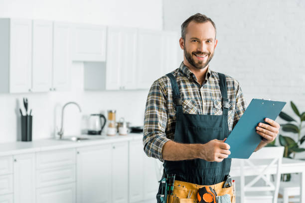 smiling handsome plumber holding clipboard and looking at camera in kitchen smiling handsome plumber holding clipboard and looking at camera in kitchen technician photos stock pictures, royalty-free photos & images