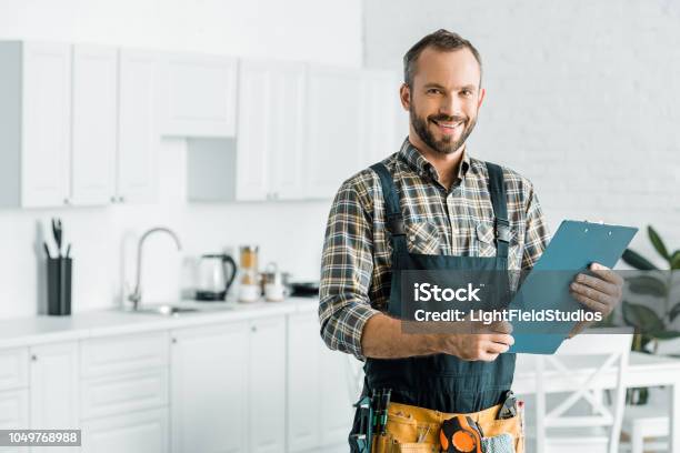 Smiling Handsome Plumber Holding Clipboard And Looking At Camera In Kitchen Stock Photo - Download Image Now