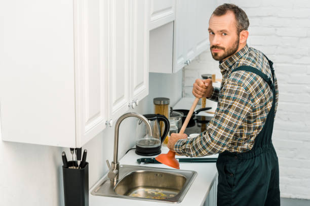 istock handsome plumber using plunger and cleaning sink in kitchen, looking at camera 1049769044