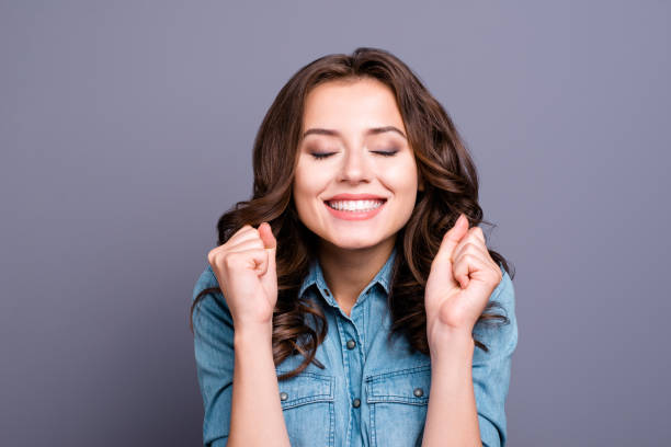moda dulce tierna magnífico alegre adorable encantador atractivo elegante morena chica con pelo ondulado en camisa casual mezclilla, mostrando el gesto de la suerte, cerró ojos, aislados sobre fondo gris - oracion para ganar loteria fotografías e imágenes de stock