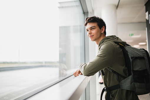 A young confident man looking out of a window, he is wearing a large backpack and casual clothing.