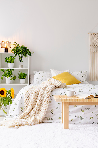 Sunny spring bedroom interior with green plants beside a bed dressed in eco cotton linen. Yellow accents. Empty white background wall. Real photo.