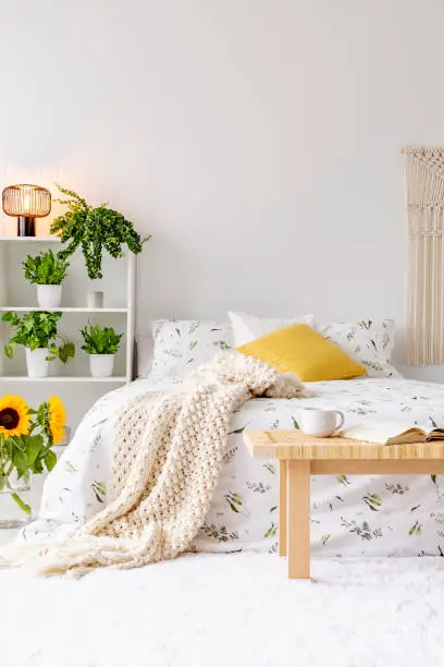 Photo of Sunny spring bedroom interior with green plants beside a bed dressed in eco cotton linen. Yellow accents. Empty white background wall. Real photo.