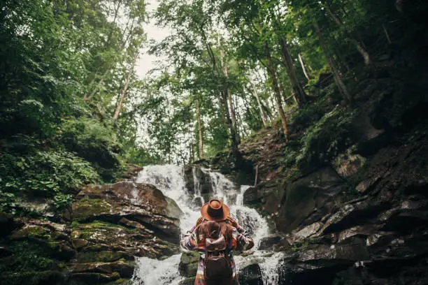 Photo of stylish hipster girl in hat with backpack looking at waterfall in forest in mountains. traveler woman exploring woods. travel and wanderlust concept. atmospheric moment