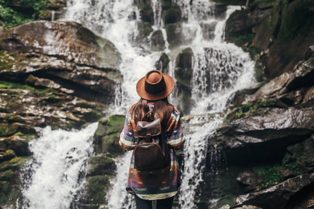 istock stylish hipster girl in hat with backpack looking at waterfall in forest in mountains. traveler woman exploring woods. travel and wanderlust concept. summer vacation, space for text 1049836202