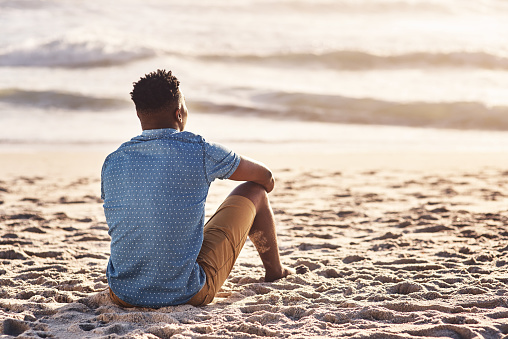 Rearview shot of a young man sitting at the water's edge and looking at the view of the ocean