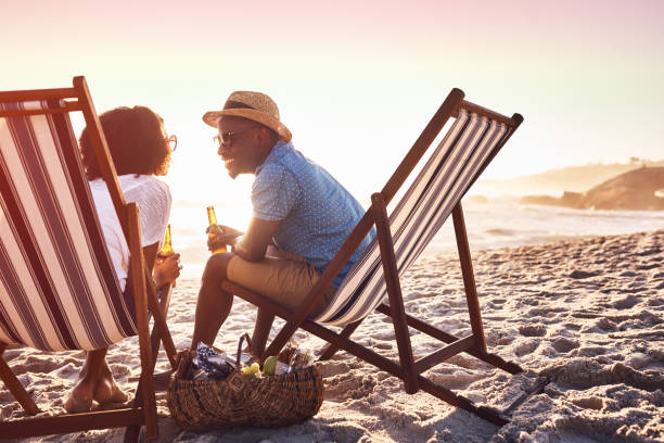 It wouldn't be summer without a bit of romance Shot of a happy young couple relaxing on chairs and having drinks at sunset on the beach black couple sunset stock pictures, royalty-free photos & images
