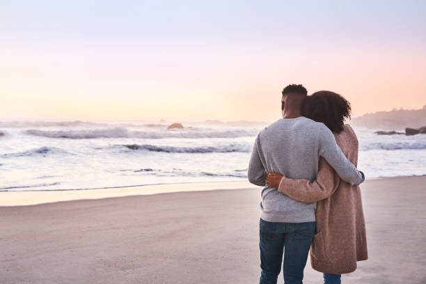 The sunset seems closer at the beach Rearview shot of a happy young couple standing by the water’s edge and looking at the view of the ocean falling in love photos stock pictures, royalty-free photos & images