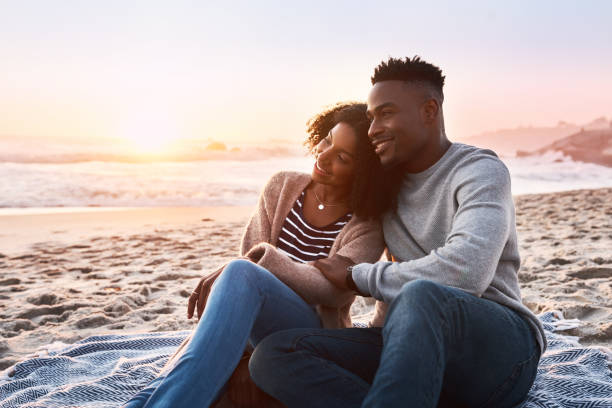 Live where the sun meets the ocean Shot of a happy young couple relaxing on a blanket during sunset at the beach black couple sunset stock pictures, royalty-free photos & images