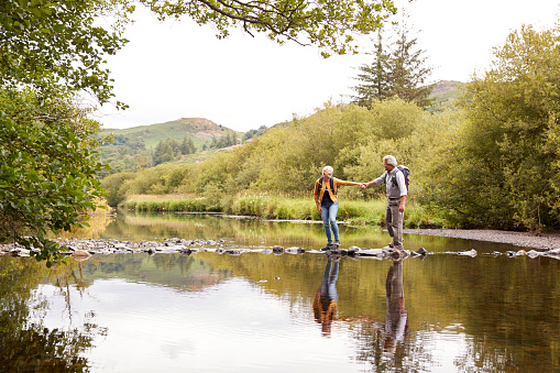 Senior Couple Crossing River Whilst Hiking In UK Lake District