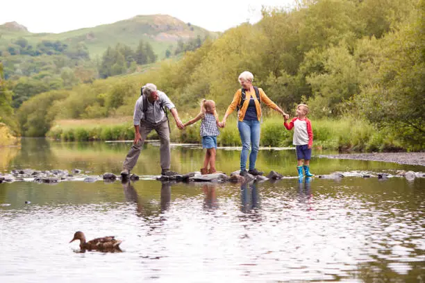 Photo of Grandparents With Grandchildren Crossing River Whilst Hiking In UK Lake District