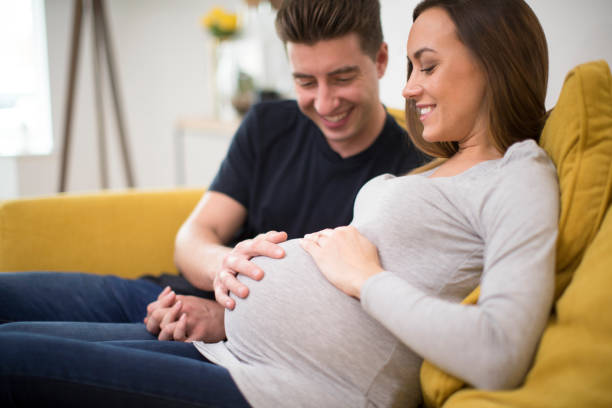 coppia incinta seduta sul divano con l'uomo che tocca lo stomaco delle donne - domestic life family heterosexual couple young family foto e immagini stock