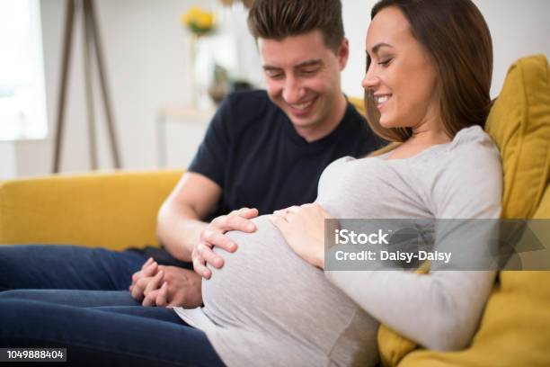 Pregnant Couple Sitting On Sofa With Man Touching Womans Stomach Stock Photo - Download Image Now