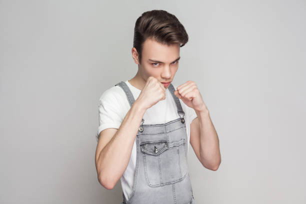 retrato de grave morena joven de estilo casual con mono de camiseta y denim de pie y mirando a cámara boxeo puños y listo para atacar. - serious punching fighting displeased fotografías e imágenes de stock