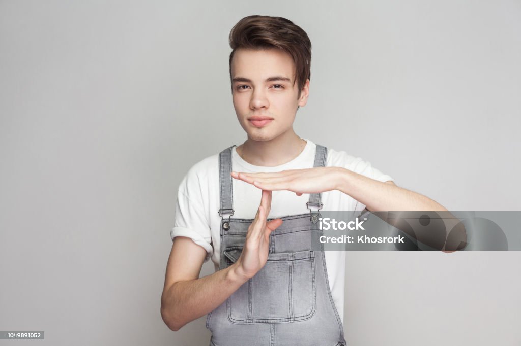 Portrait of serious young brunette man in casual style with denim overalls looking at camera, showing timeout gesture and asking for more few time Portrait of serious young brunette man in casual style with denim overalls looking at camera, showing timeout gesture and asking for more few time. indoor studio shot, isolated on gray background. Teenage Boys Stock Photo