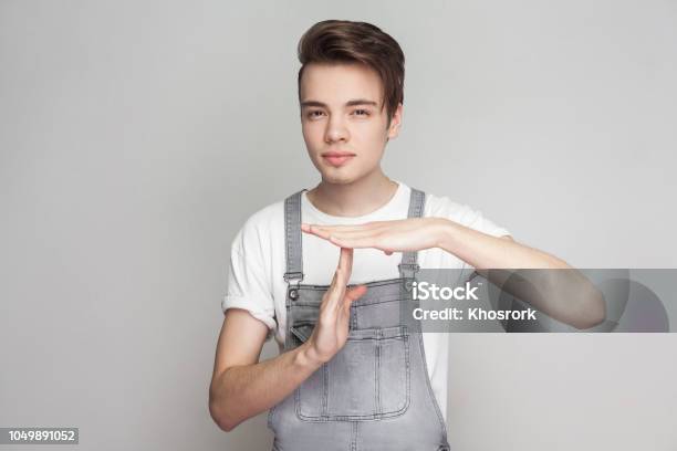Portrait Of Serious Young Brunette Man In Casual Style With Denim Overalls Looking At Camera Showing Timeout Gesture And Asking For More Few Time Stock Photo - Download Image Now