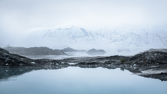 The icefall of the Matanuska Glacier is viewed through a thick morning fog. In the foreground, a large kettle pond as well as mounds of dirt and rock moraine from the glacier are visible.