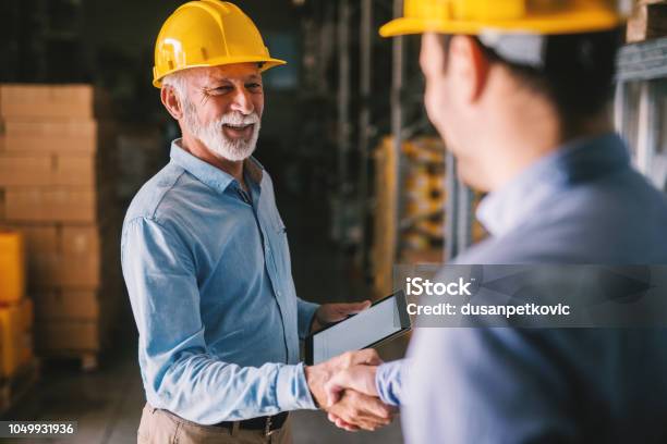 Two Satisfied Business Man Standing In Warehouse With Helmets On Their Head Shaking Hands And Closing Good Job Cooperation Stock Photo - Download Image Now