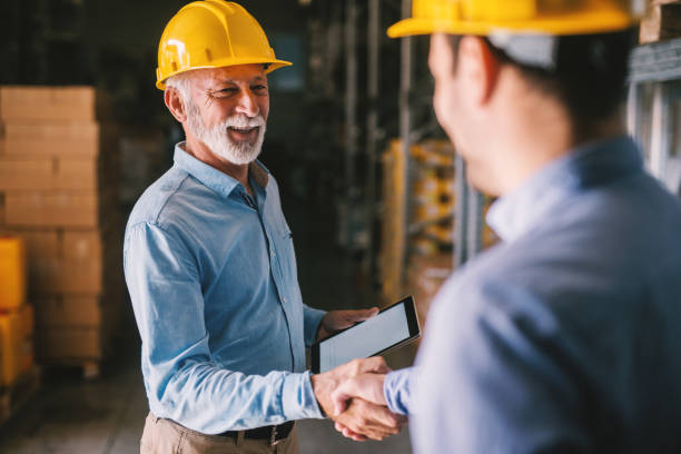 two satisfied business man standing in warehouse with helmets on their head. shaking hands and closing good job cooperation. - manual worker handshake industry warehouse imagens e fotografias de stock